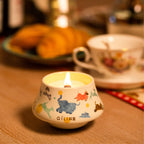 Scented candle in illustrated ceramic cup with visible flame on wood